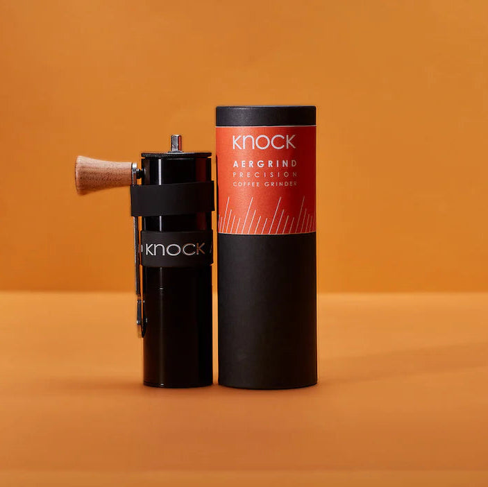 A compact AERGRIND hand coffee grinder with a one-piece aluminium upper body and walnut wood knob, displayed next to its packaging.